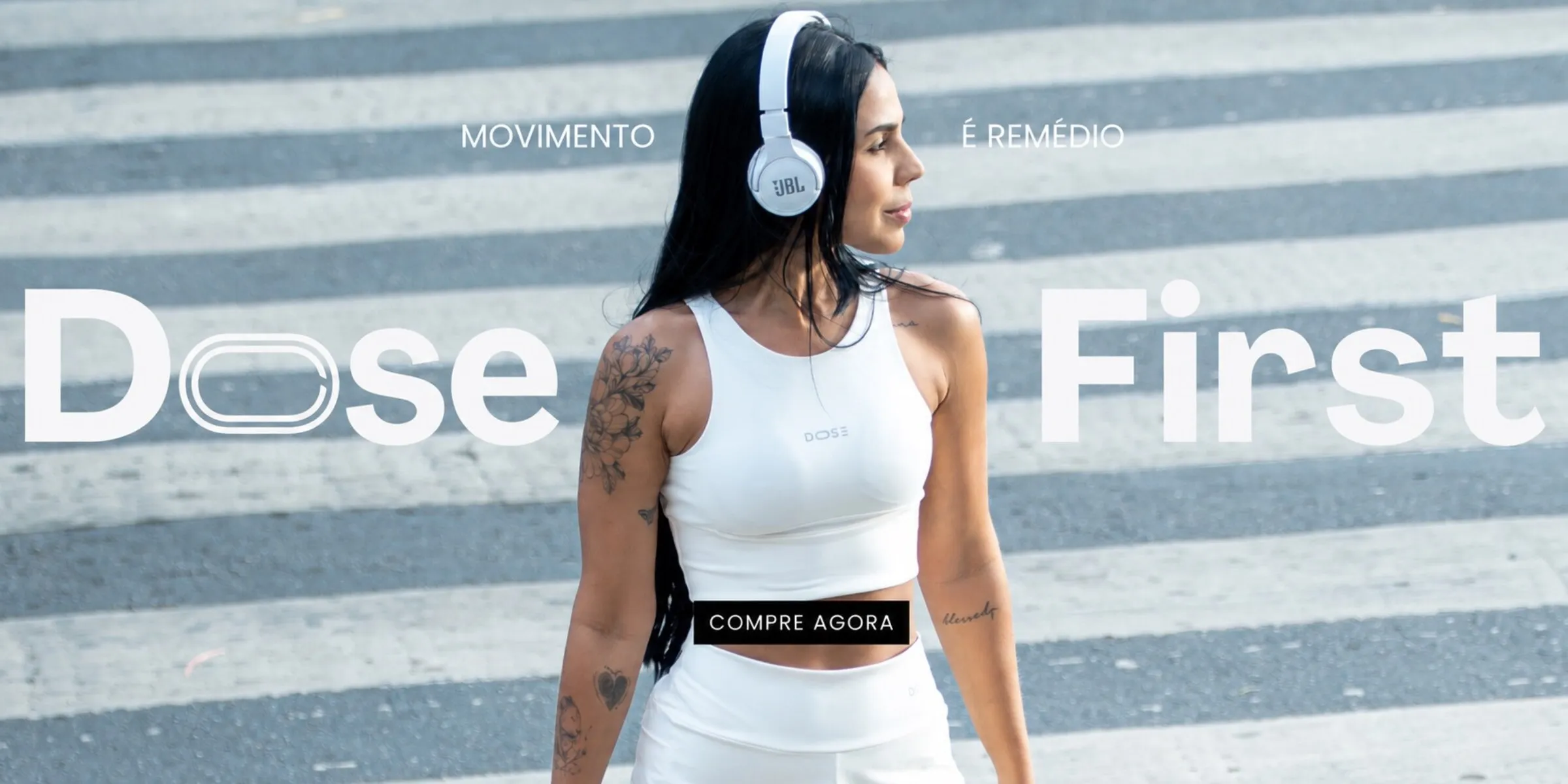 Movimento é remédio. Dose First — modelo com o Conjunto Run DOSE claro e fones de ouvido, atravessando uma faixa de pedestres. Compre agora.